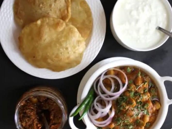 Two puffed bhature with a bowl of chole, sliced onion, yoghurt and pickle